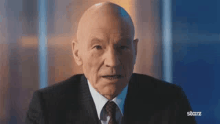 A Bald Man In A Suit And Tie Is Sitting In Front Of A Blue Wall . GIF