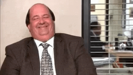 A Bald Man In A Suit And Tie Is Smiling While Sitting In Front Of A Window In An Office . GIF