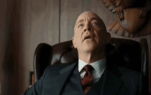 A Bald Man In A Suit And Tie Sits In A Chair GIF