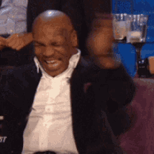 A Bald Man In A Suit And White Shirt Is Sitting In A Chair With His Fist In The Air . GIF