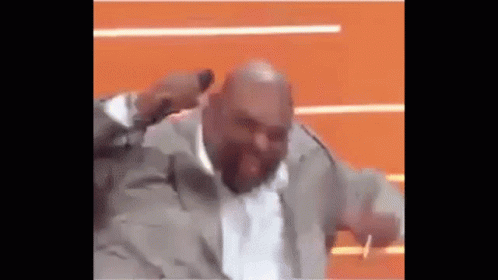 A Bald Man In A Suit Is Dancing On A Stage With His Hands In The Air . GIF