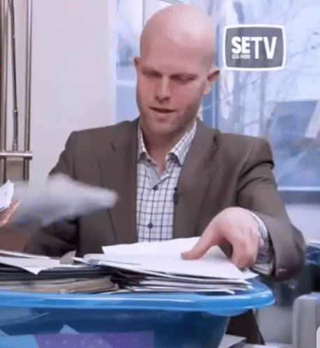 A Bald Man In A Suit Is Sitting At A Desk With Papers In A Blue Bin . GIF