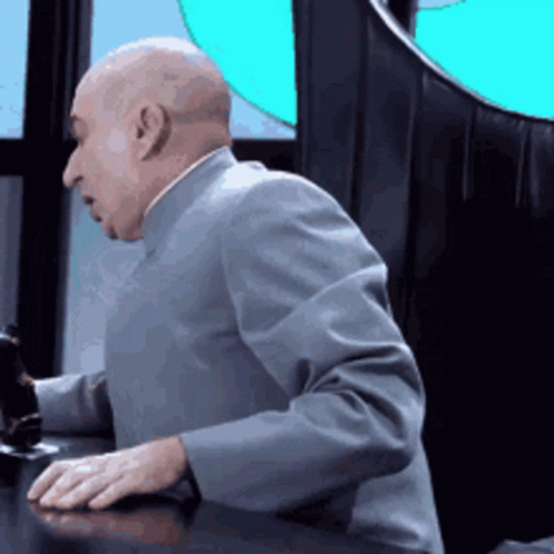A Bald Man In A Suit Is Sitting At A Table With His Hands On The Desk . GIF