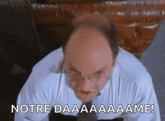 A Bald Man In A White Shirt And Tie Is Sitting In Front Of A Table And Making A Funny Face . GIF
