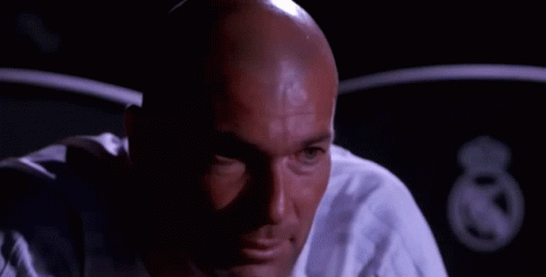 A Bald Man In A White Shirt Is Smiling And Looking At The Camera In A Dark Room . GIF