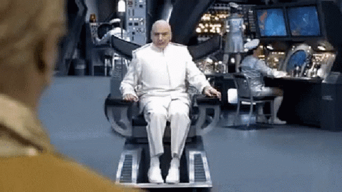 A Bald Man In A White Suit Is Sitting On A Chair In A Room . GIF
