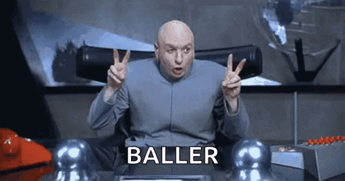 A Bald Man Is Giving A Peace Sign While Sitting At A Desk With The Word Baller Written On It . GIF