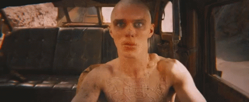 A Bald Man Is Holding A Gun In A Car With Flames Coming Out Of The Back Seat . GIF