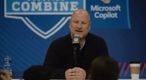a bald man is speaking into a microphone in front of a sign that says combine