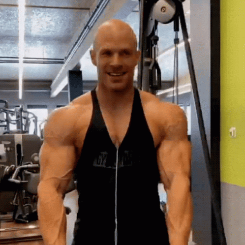 A Bald Man Is Standing In A Gym Wearing Headphones And A Black Tank Top . GIF