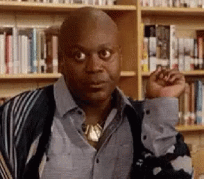 A Bald Man Is Standing In Front Of A Bookshelf In A Library With His Fist In The Air . GIF