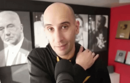 A Bald Man Is Standing In Front Of A Microphone In A Room With Pictures On The Wall Behind Him . GIF
