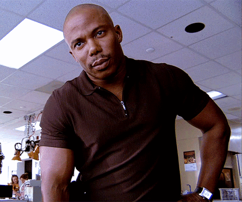 A Bald Man Wearing A Brown Polo Shirt And A Watch Stands In A Room GIF