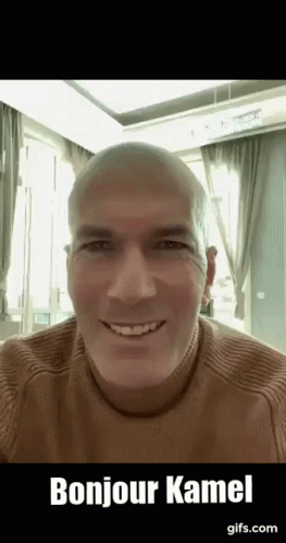 A Bald Man Wearing A Brown Sweater Is Smiling And Saying Bonjour Kamel . GIF