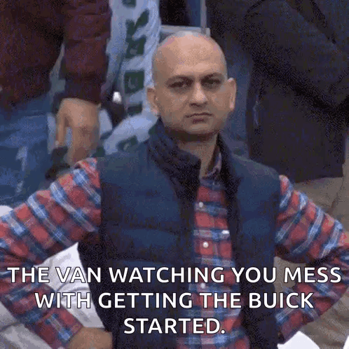 A Bald Man Wearing A Plaid Shirt And Vest Is Standing In A Crowd . GIF