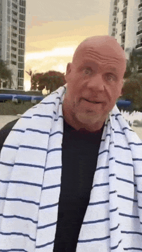 A Bald Man Wearing A Striped Towel Around His Neck Is Standing In Front Of A Building . GIF