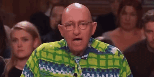 A Bald Man Wearing Glasses And A Green And Blue Shirt Is Speaking In Front Of A Crowd . GIF