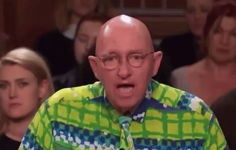 A Bald Man Wearing Glasses And A Green And Blue Shirt Is Speaking In Front Of A Crowd . GIF