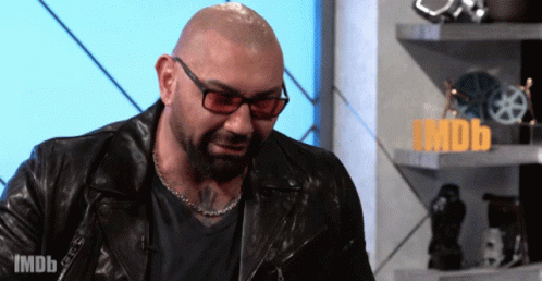 A Bald Man Wearing Glasses And A Leather Jacket Stands In Front Of A Sign That Says Imdb On It GIF