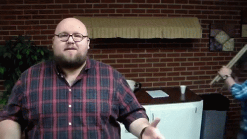 A Bald Man Wearing Glasses And A Plaid Shirt Stands In Front Of A Brick Wall GIF