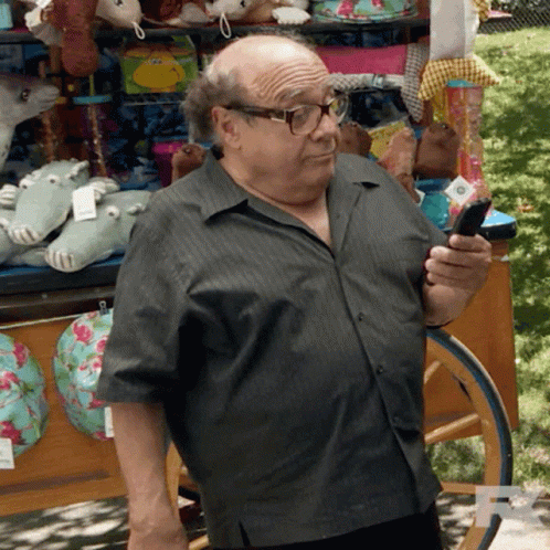 A Bald Man Wearing Glasses Is Looking At His Phone In Front Of A Cart Full Of Stuffed Animals GIF