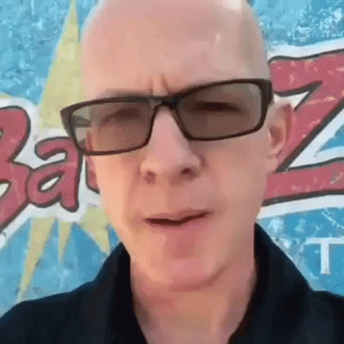 A Bald Man Wearing Sunglasses And A Black Shirt Stands In Front Of A Wall With The Letter Z On It GIF