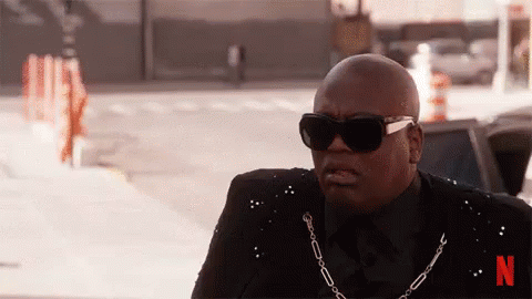 A Bald Man Wearing Sunglasses And A Chain Around His Neck Is Sitting On The Sidewalk . GIF