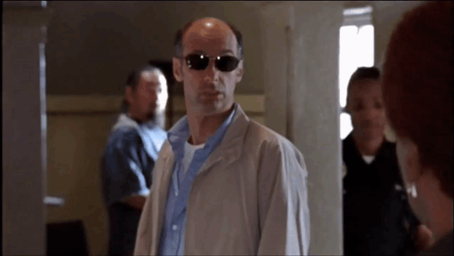 A Bald Man Wearing Sunglasses And A Jacket Stands In A Hallway GIF