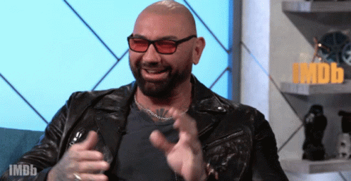 A Bald Man Wearing Sunglasses And A Leather Jacket Is Sitting In Front Of An Imdb Sign GIF