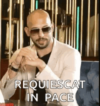 A Bald Man Wearing Sunglasses And A Suit Is Sitting At A Table With His Hands Folded . GIF