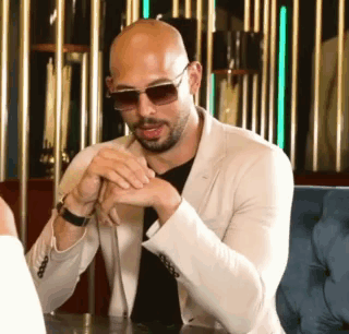 A Bald Man Wearing Sunglasses And A White Jacket Is Sitting At A Table In A Restaurant . GIF
