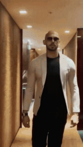 A Bald Man Wearing Sunglasses And A White Jacket Walks Down A Hallway . GIF