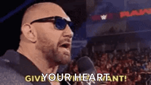 A Bald Man Wearing Sunglasses Is Standing In Front Of A Microphone And Saying `` Give Your Heart ! '' GIF
