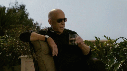A Bald Man Wearing Sunglasses Sits On A Chair Holding A Glass GIF