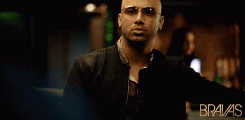 A Bald Man Wearing Sunglasses Stands In Front Of An Ad For Bravas GIF