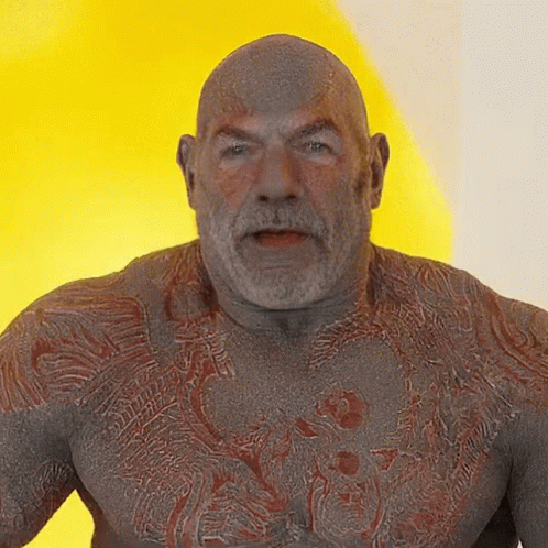 A Bald Man With A Beard And A Tattoo On His Chest Looks At The Camera GIF