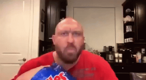 A Bald Man With A Beard Is Eating A Bag Of Potato Chips In A Kitchen . GIF