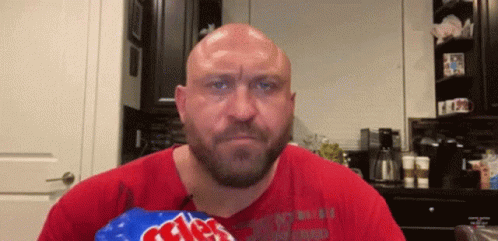 A Bald Man With A Beard Is Holding A Bag Of Chips In His Hand . GIF