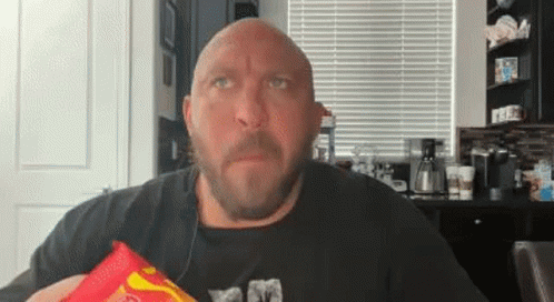 A Bald Man With A Beard Is Holding A Bag Of Chips In His Hand . GIF