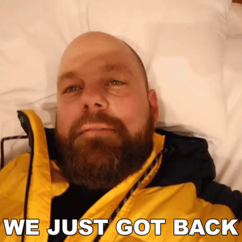 A Bald Man With A Beard Is Laying On A Bed With The Words " We Just Got Back " Above Him GIF