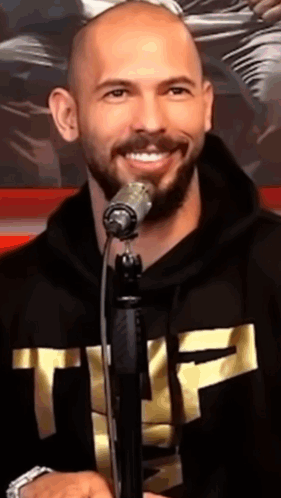 A Bald Man With A Beard Is Singing Into A Microphone While Wearing A Black Hoodie With The Letter T On It GIF