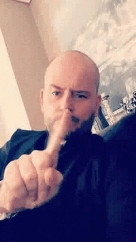 A Bald Man With A Beard Is Sitting On A Couch Making A Rude Gesture With His Middle Finger . GIF