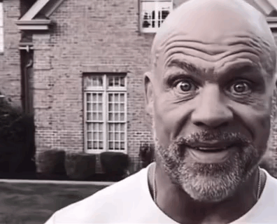 A Bald Man With A Beard Is Smiling In Front Of A Brick Building . GIF