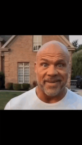 A Bald Man With A Beard Is Standing In Front Of A Brick House . GIF