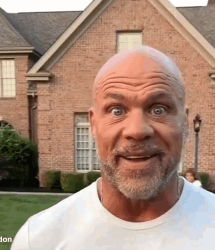 A Bald Man With A Beard Is Standing In Front Of A Large Brick House . GIF