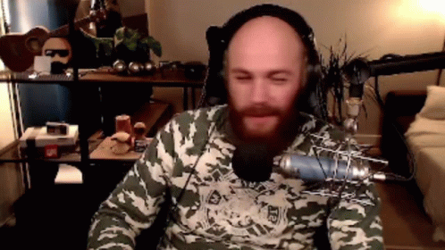 A Bald Man With A Beard Is Wearing Headphones And A Camo Shirt While Sitting In Front Of A Microphone . GIF