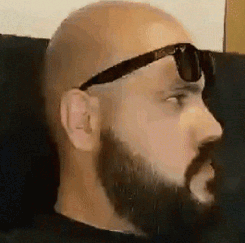A Bald Man With A Beard Is Wearing Sunglasses While Sitting On A Couch . GIF