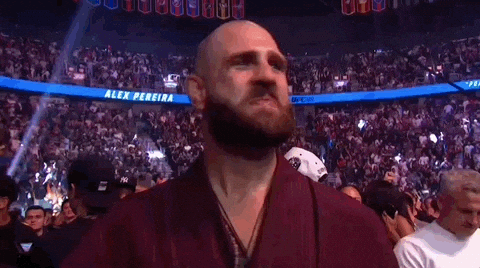 A Bald Man With A Beard Stands In Front Of A Crowd In A Stadium With A Banner That Says Alex Pereira . GIF