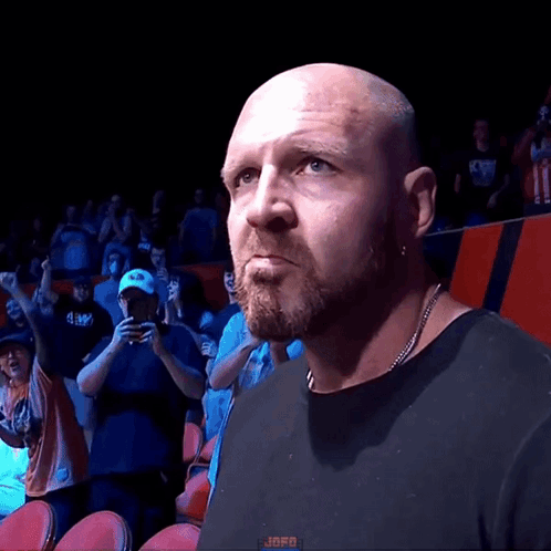 A Bald Man With A Beard Wearing A Black Shirt Stands In Front Of A Crowd With The Word Jafo On The Bottom Left GIF