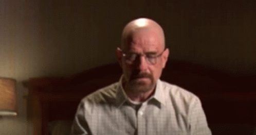 A Bald Man With A Beard Wearing Glasses Is Sitting In Front Of A Bed GIF